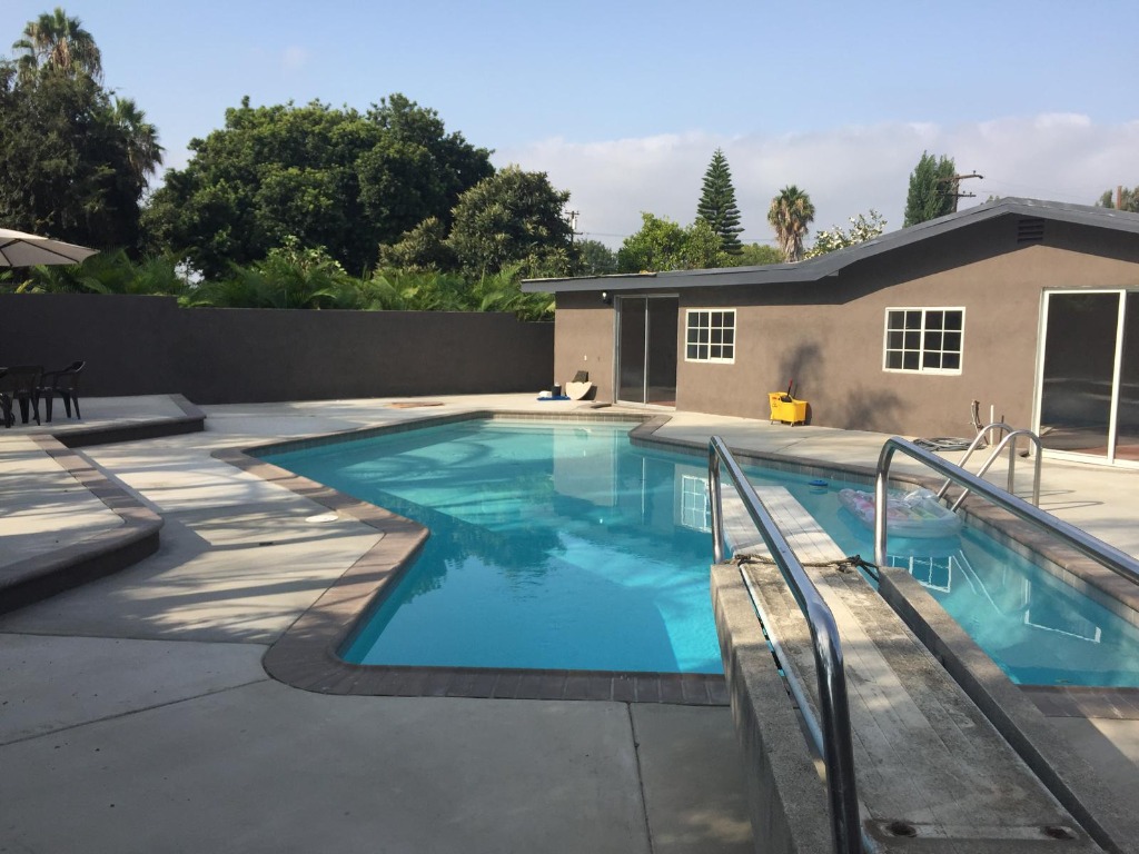 Completed inground pool remodel with crystal clear turquoise water, concrete deck, diving board, and modern dark exterior stucco